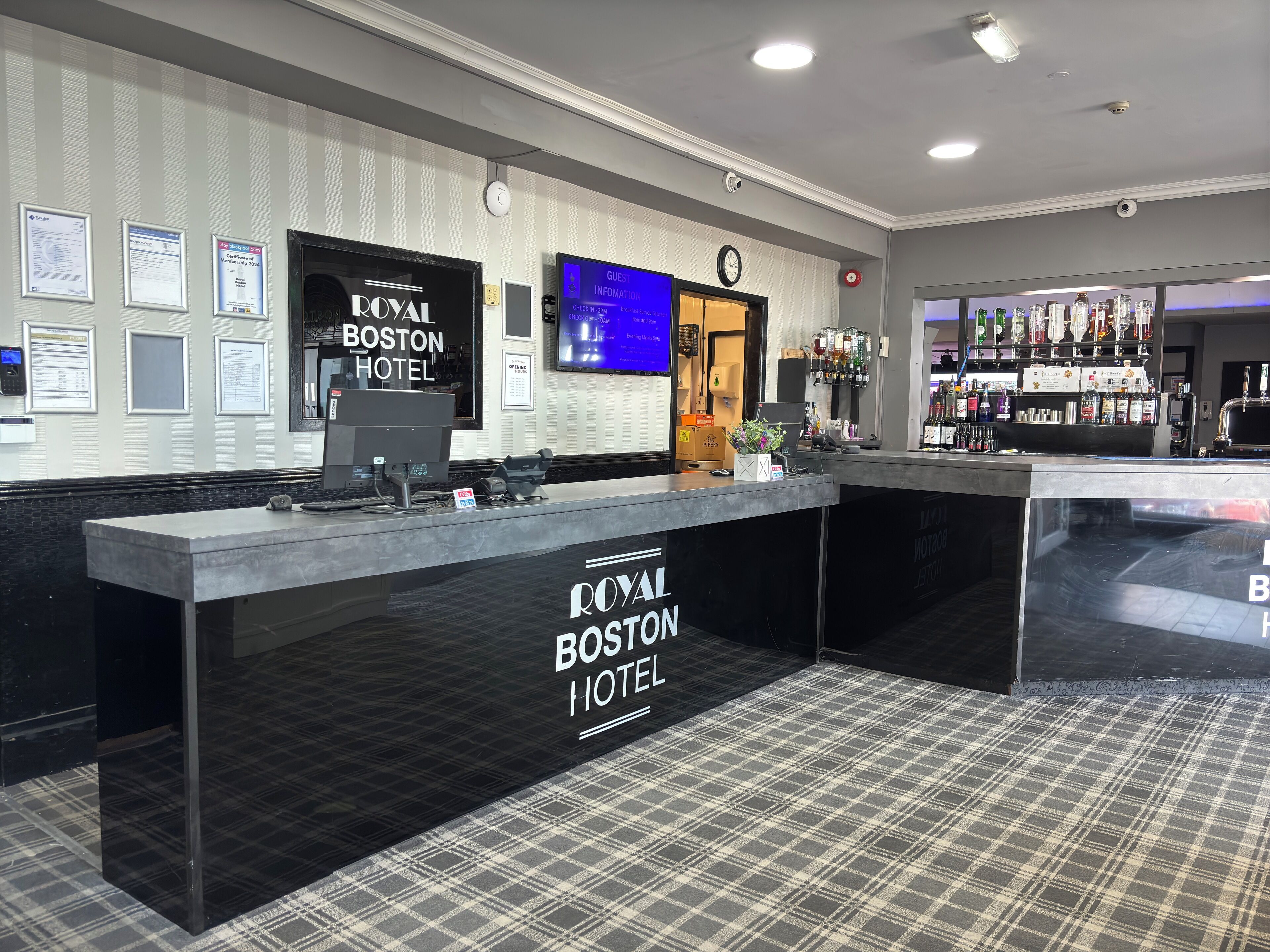 A reception desk at the Royal Boston Hotel with a check-in counter, a digital display, and a bar area with various bottles.
