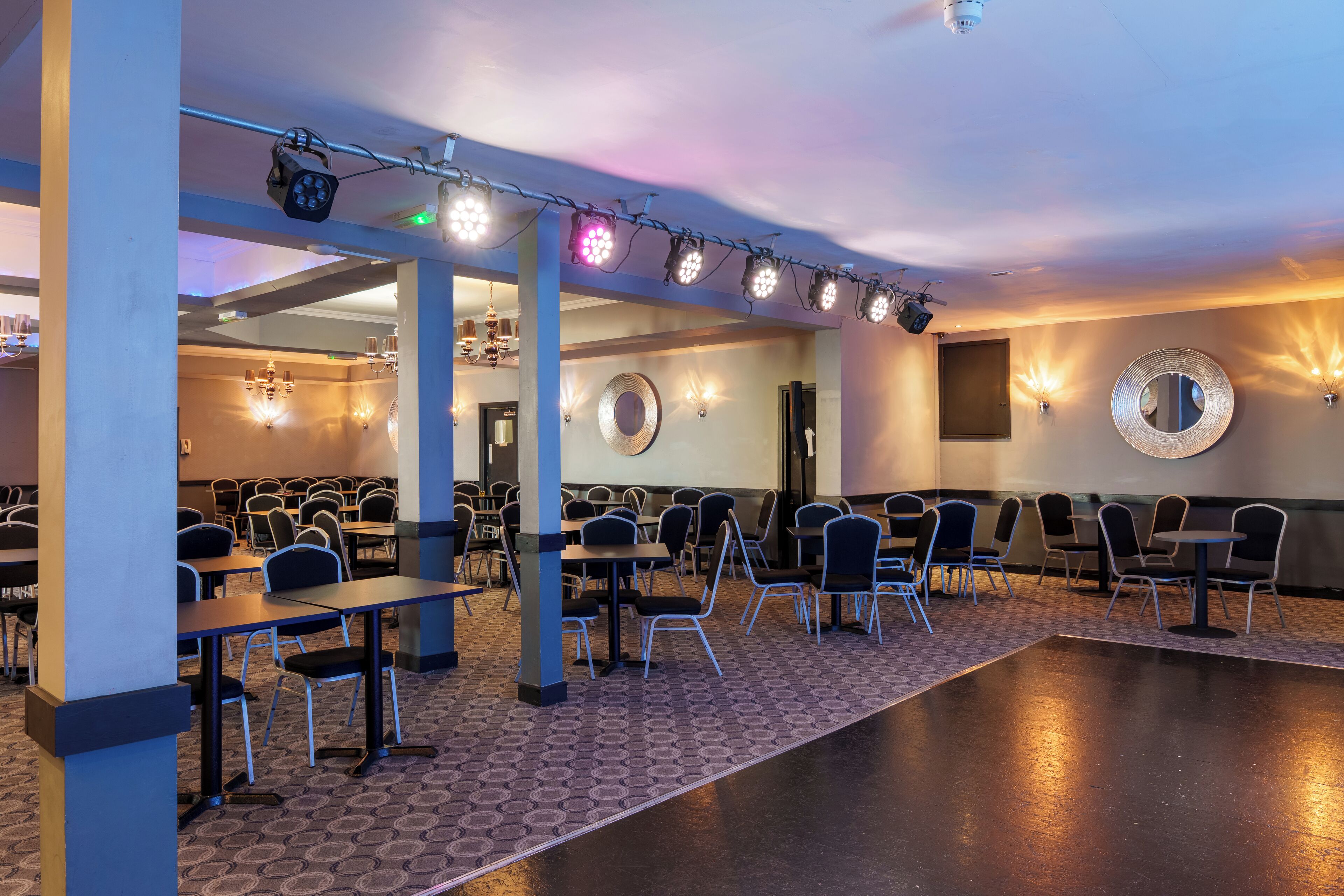 A conference room with tables and chairs, a stage with lighting, and wall sconces.