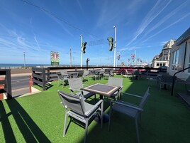 Een terras met tafels en stoelen, uitzicht op zee, en een bord met een kleurrijk ontwerp.