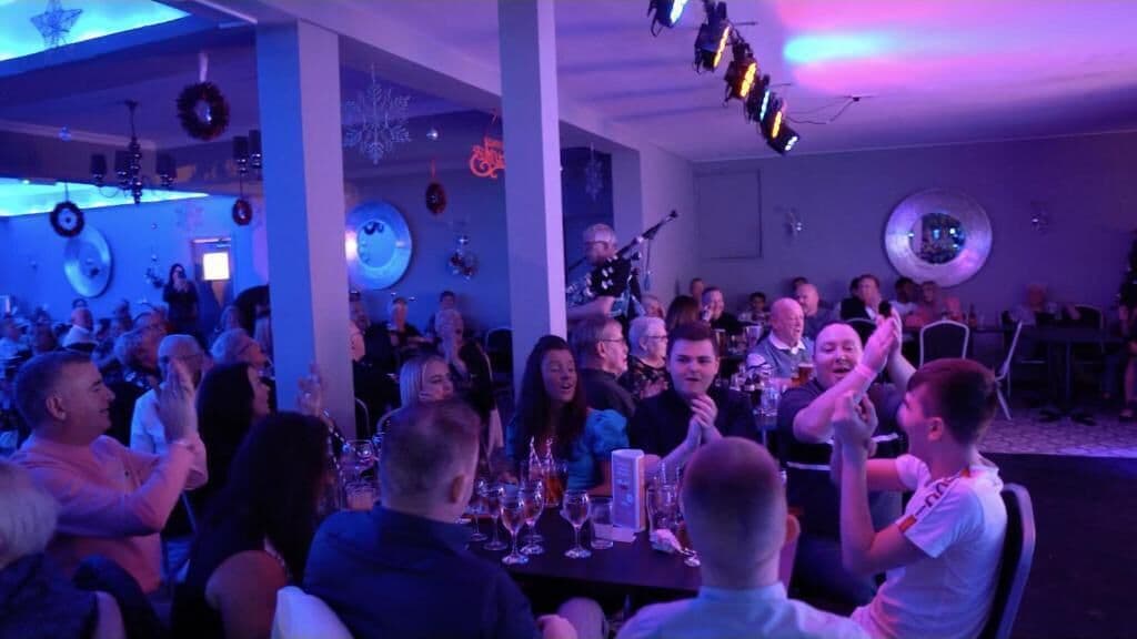 Een groep mensen zit aan een tafel in een ruimte, met een podium op de achtergrond.