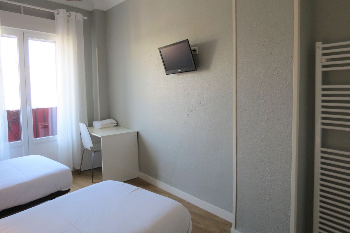 Habitación de hotel con dos camas, un escritorio, una silla, un televisor montado en la pared y una ventana con cortinas.