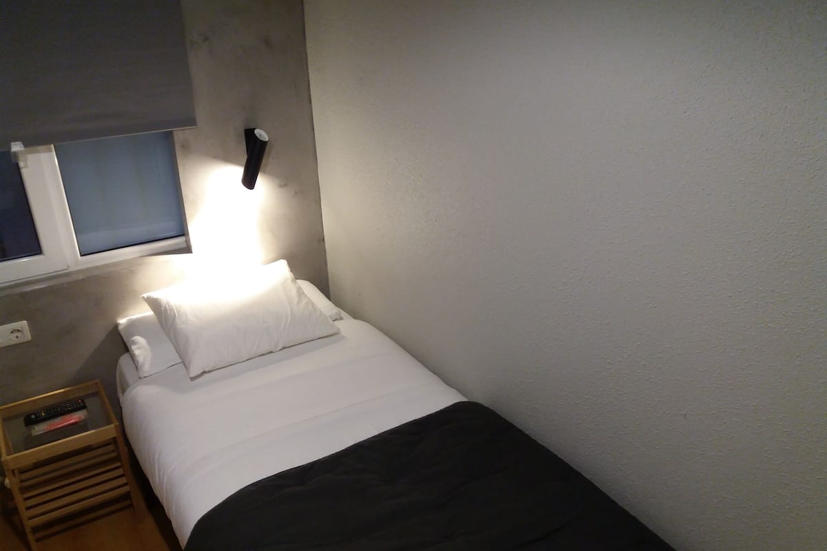 Pequeña habitación con cama individual, mesita de noche, lámpara y ventana con persianas.