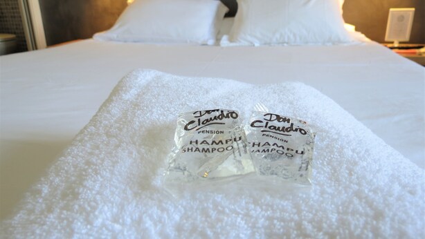 A neatly made bed with two packages of Don Claudio shampoo on a white towel. - Pension Don Claudio (Bilbao)