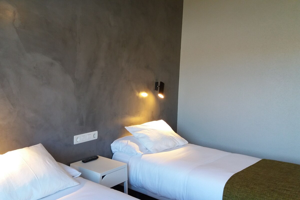 Habitación de hotel con dos camas individuales, una mesita de noche pequeña, un control remoto y una lámpara de pared.