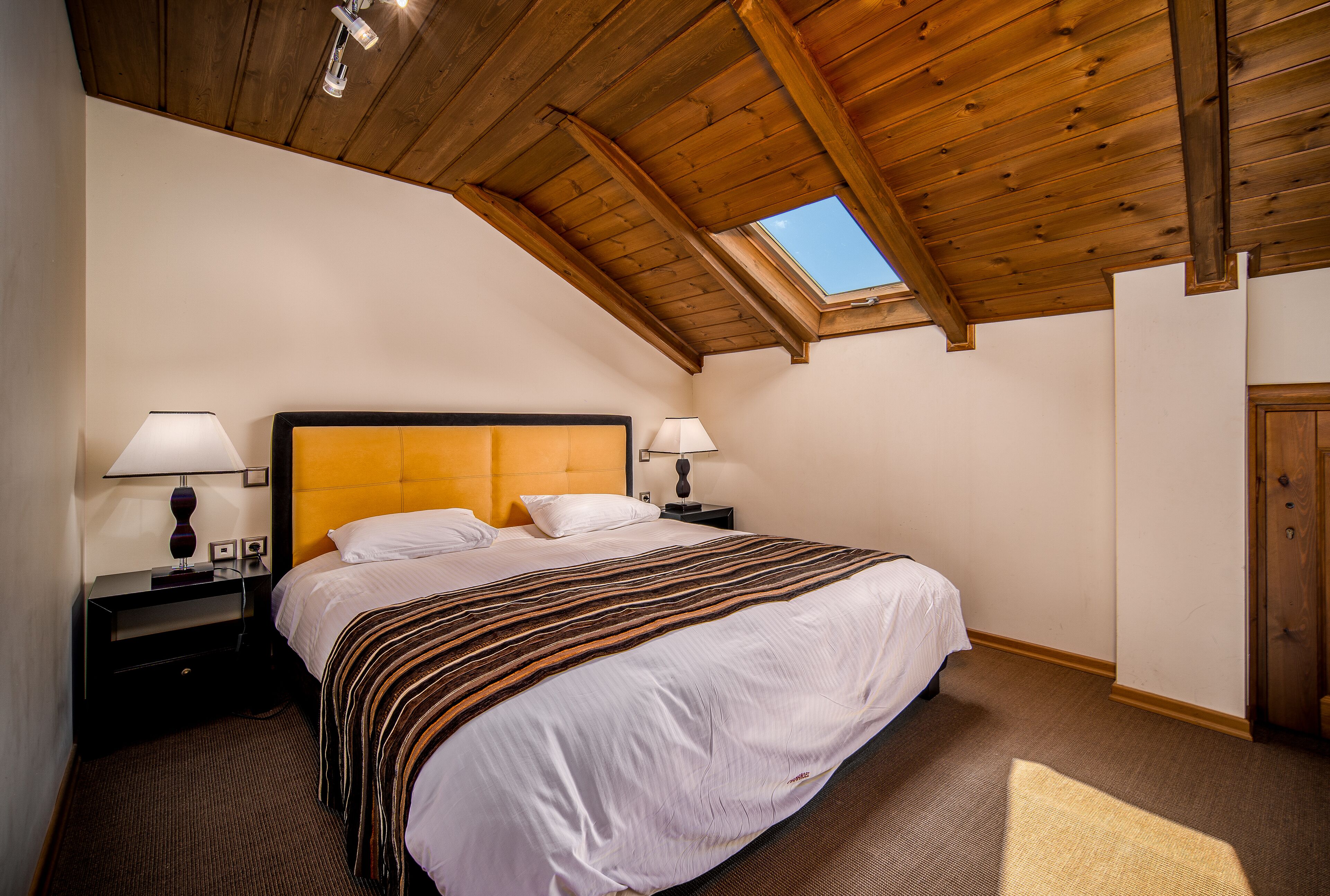 Een slaapkamer met een houten plafond, dakraam, een groot bed met wit beddengoed, twee nachtkastjes met lampen, en een houten kledingkast.