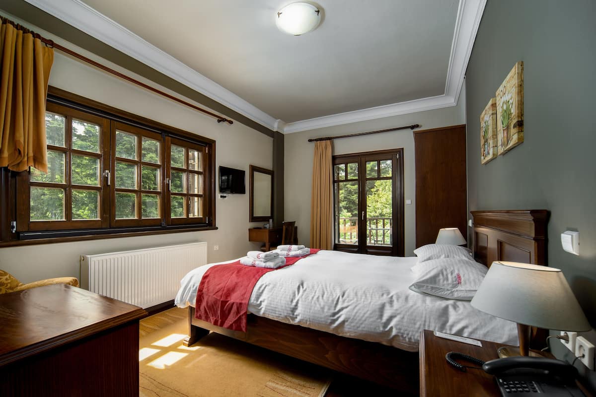 Hotellihuone, jossa on suuri sänky, työpöytä, tuoli, yöpöytä lampun kanssa ja ikkunoista avautuva näkymä vehreään maisemaan.
