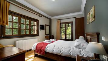 A hotel room with a large bed, a desk, a chair, a bedside table with a lamp, and a view of greenery through the windows.