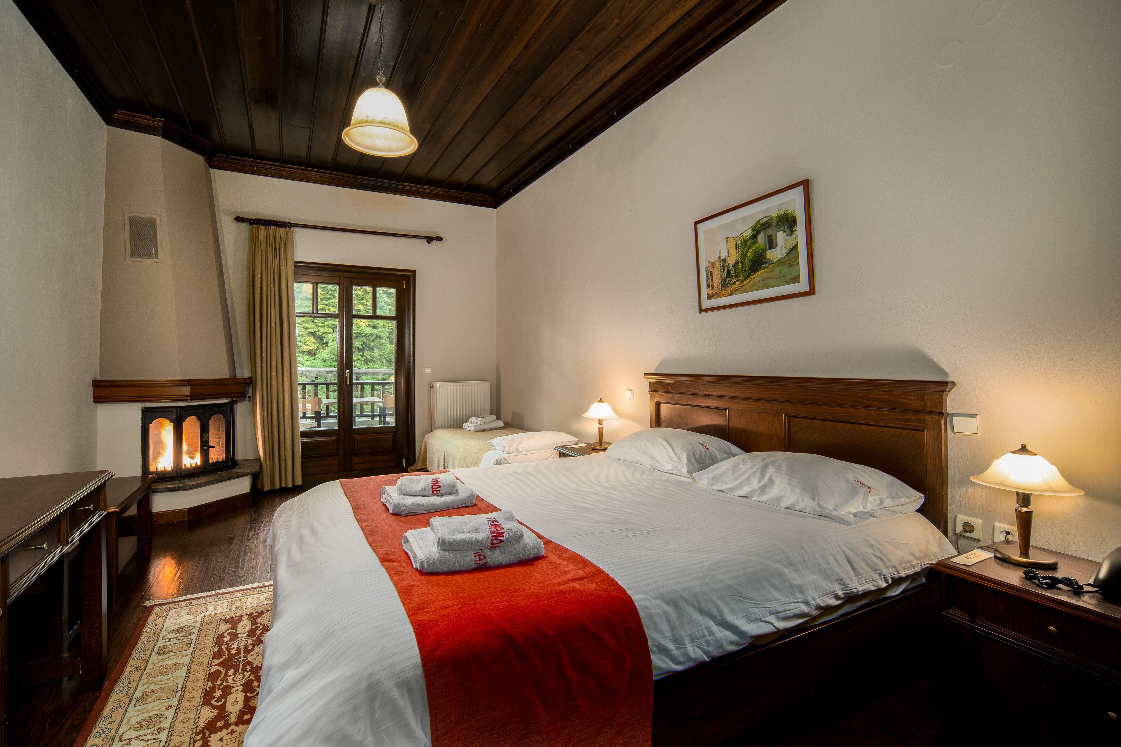 A hotel room with a large bed, a wooden headboard, a nightstand with a lamp, a fireplace, and a view of the outdoors through a window.