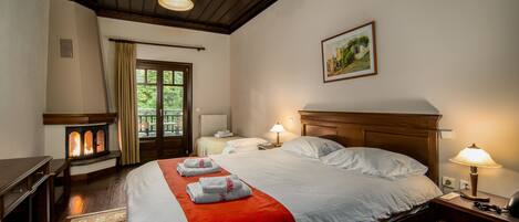 A hotel room with a large bed, a wooden headboard, a nightstand with a lamp, a fireplace, and a view of the outdoors through a window.
