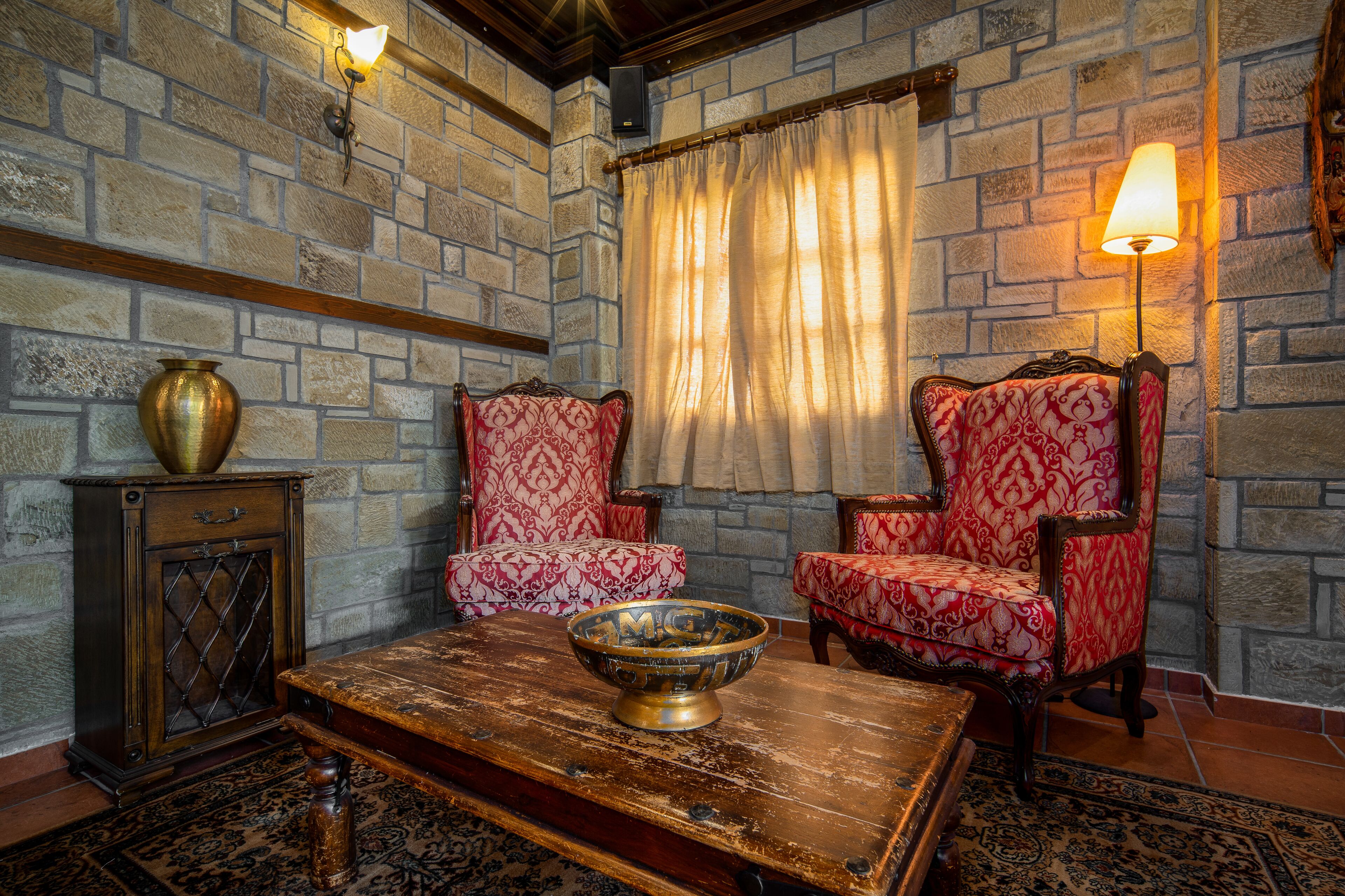 A room with stone walls, two patterned armchairs, a wooden coffee table with a bowl, and a small cabinet with a brass pot.