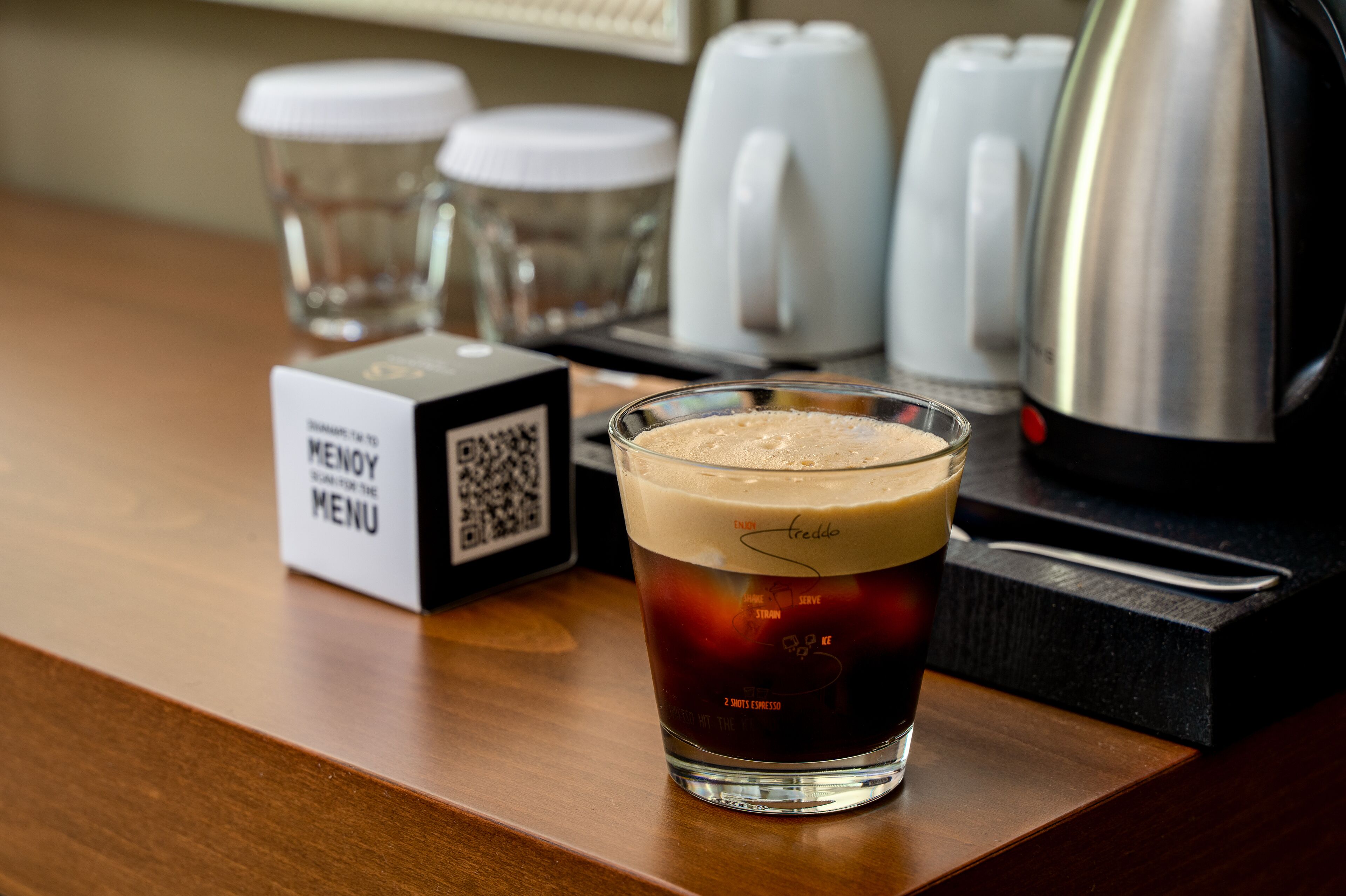 Un vaso de café helado con espuma, un cartel de menú y una cafetera sobre una mesa de madera.