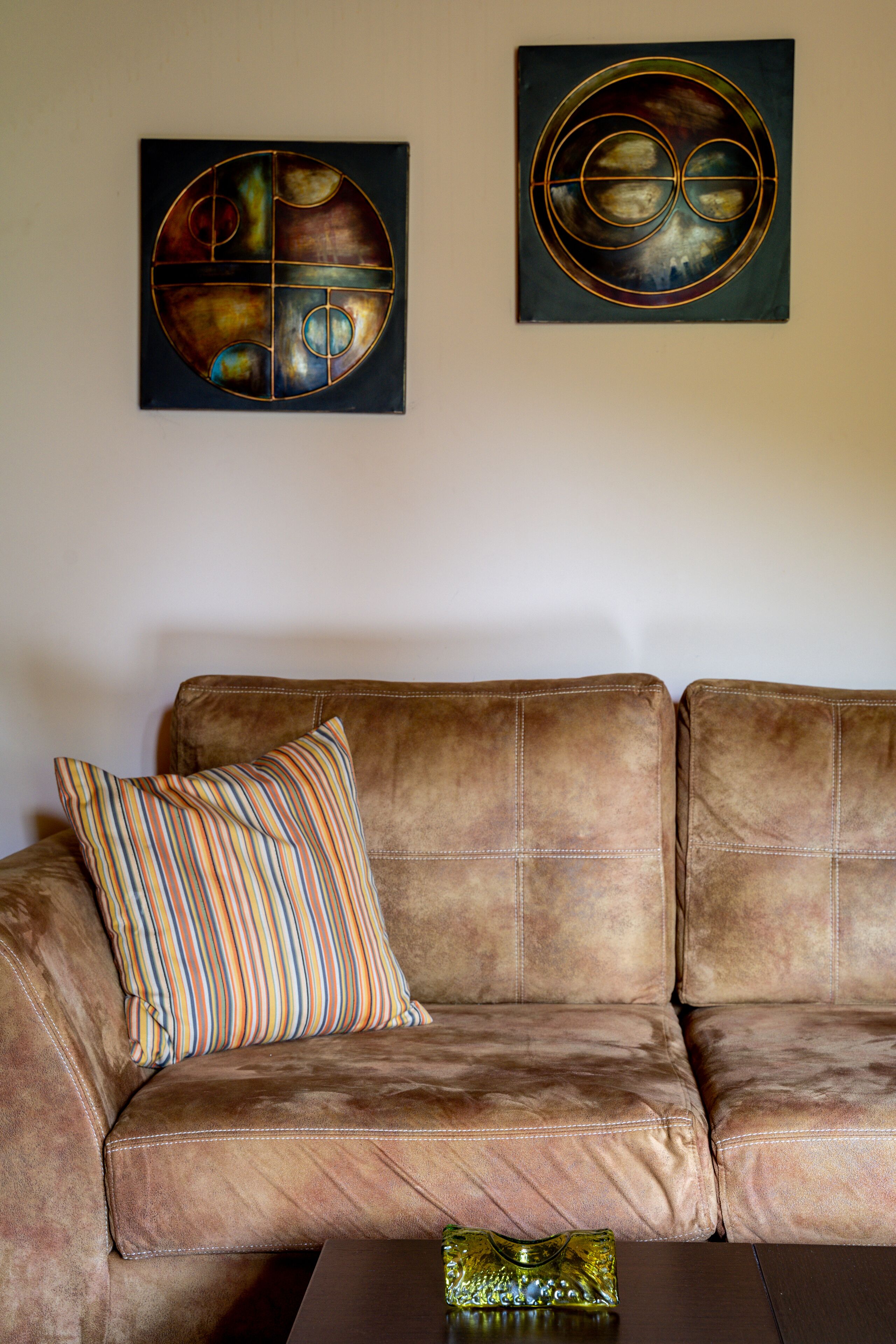A brown leather sofa with a striped pillow and a glass vase on a wooden table. Two abstract paintings are hung on the wall above the sofa.