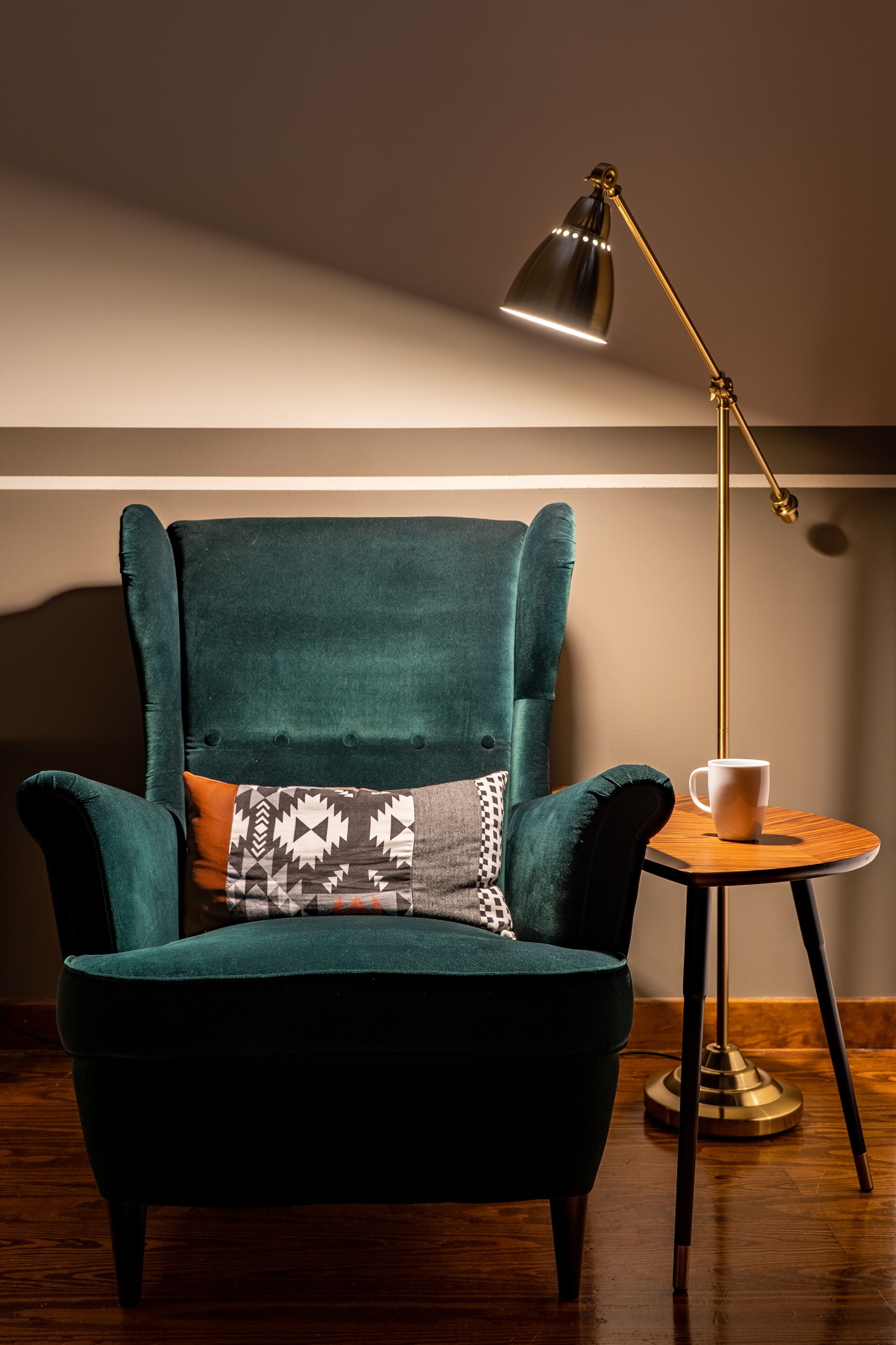 A teal armchair with a patterned cushion, a small wooden side table with a white mug, and a floor lamp with a gold base.