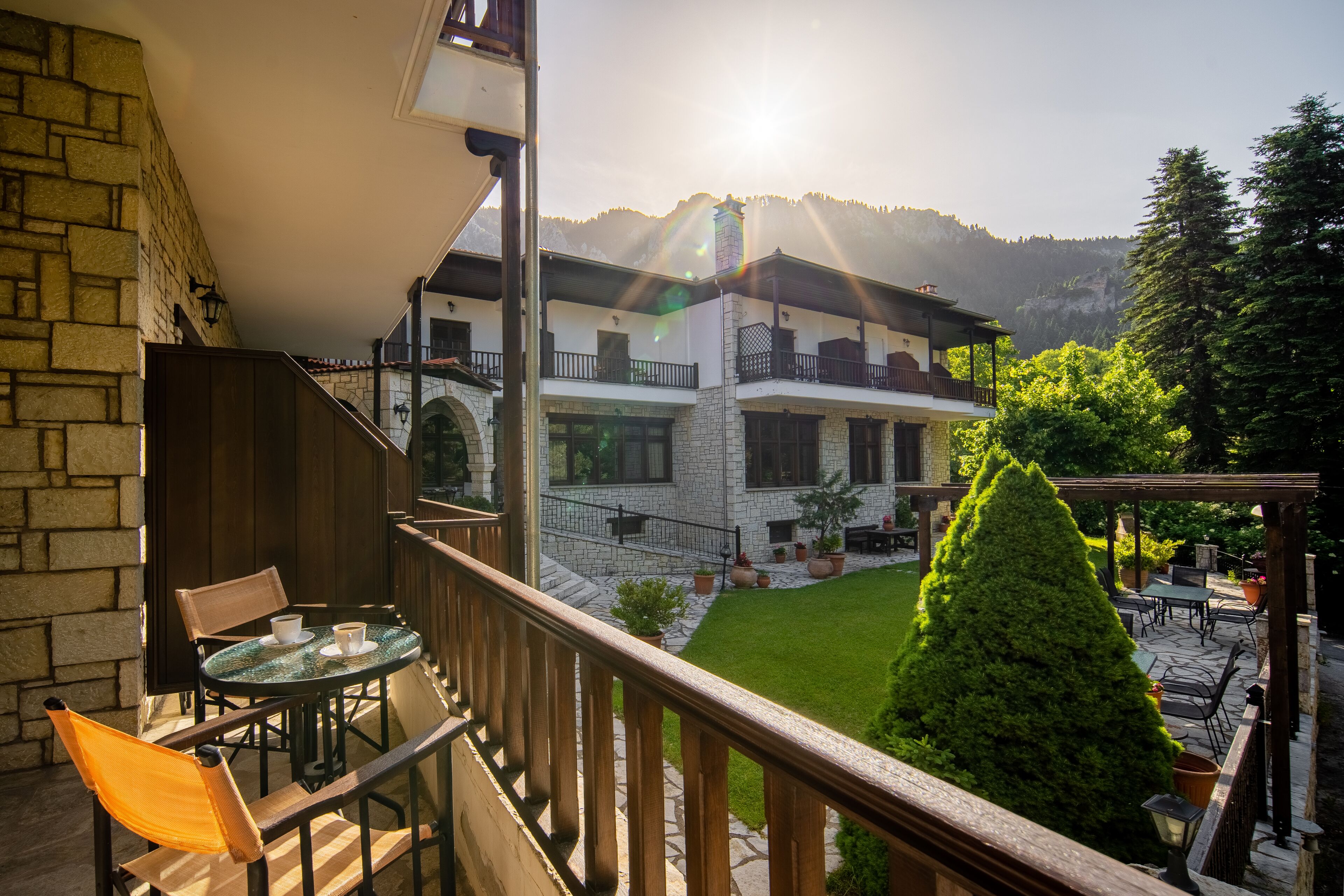 Un balcon avec une table et des chaises, un mur en pierre et une vue sur un bâtiment avec un jardin et des montagnes en arrière-plan.