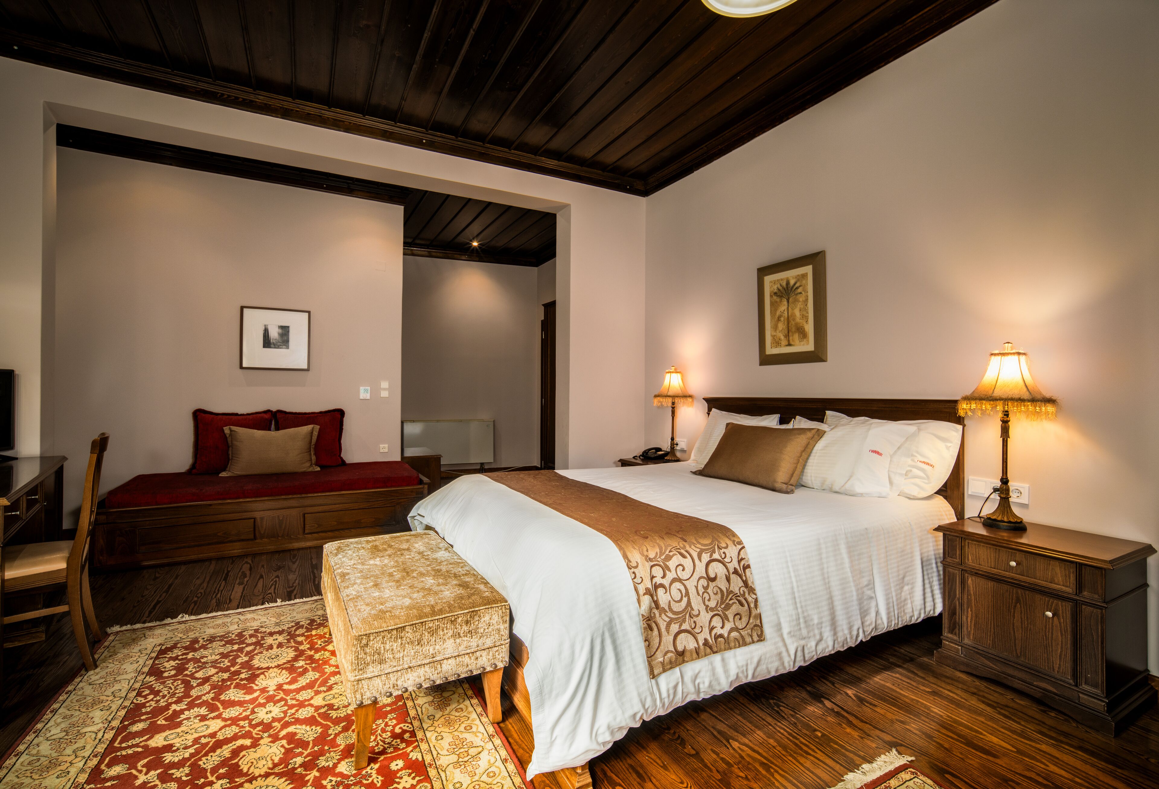 A bedroom with a large bed, two bedside lamps, a wooden nightstand, a red sofa, and a patterned rug.