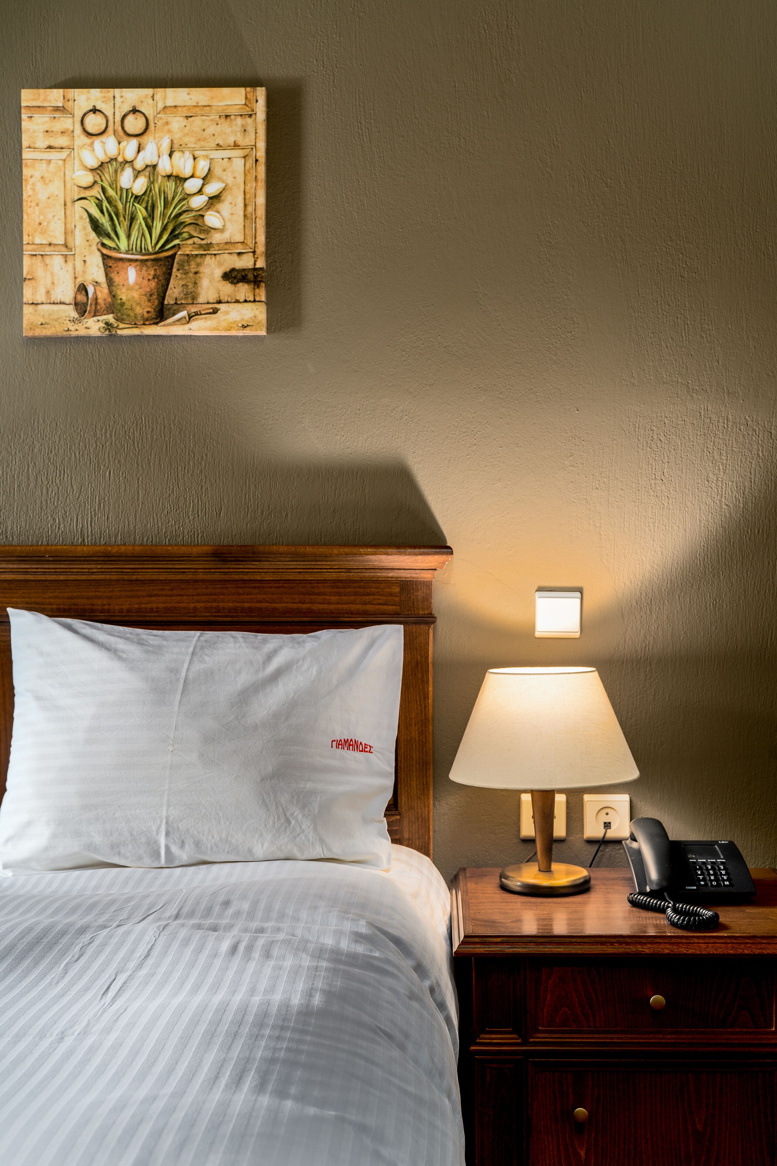 Una cama bien hecha con ropa blanca, un cabecero de madera, una lámpara de noche, un teléfono y una mesita con lámpara.