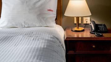 Una cama bien hecha con ropa blanca, un cabecero de madera, una lámpara de noche, un teléfono y una mesita con lámpara.