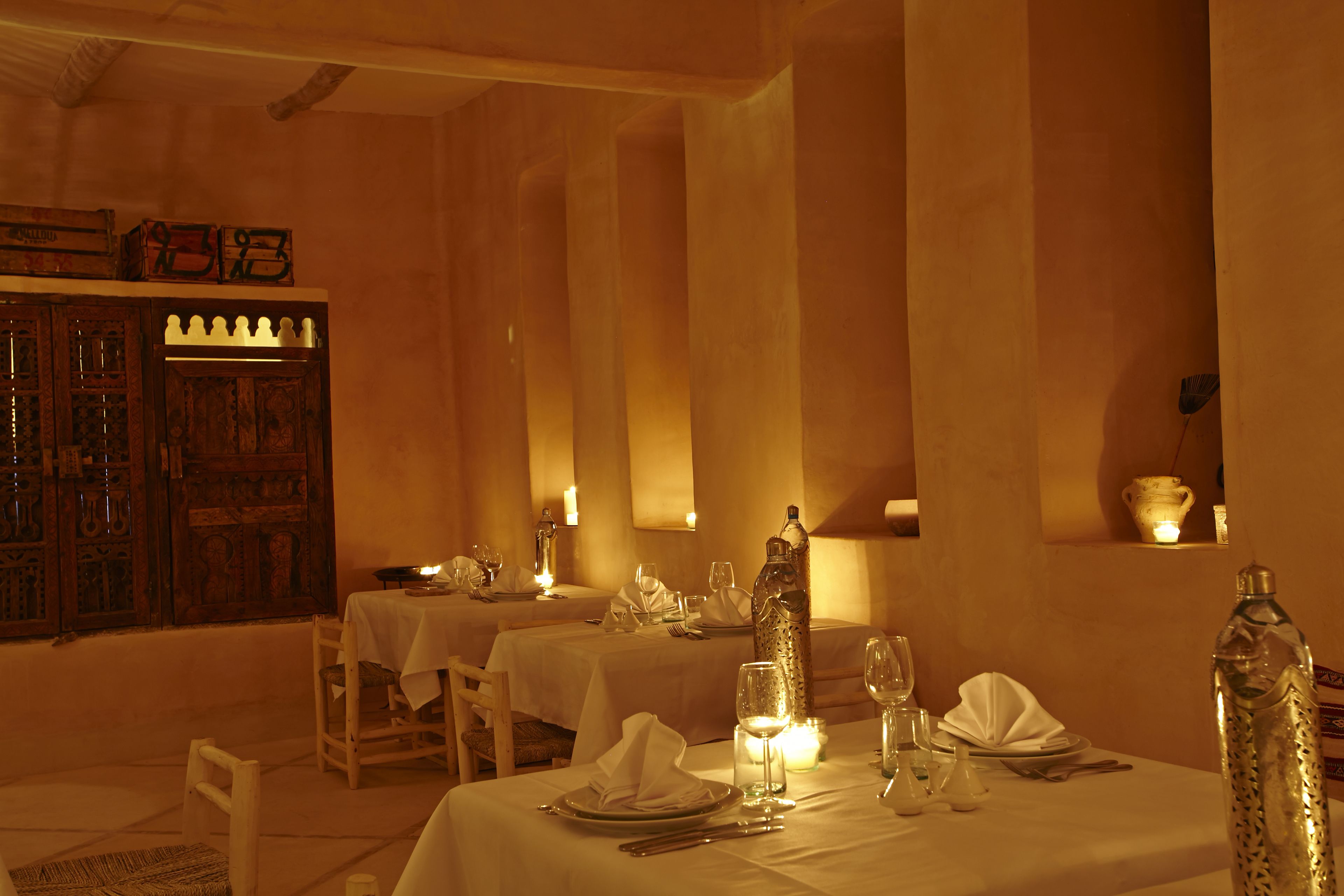 A dimly lit dining area with tables set for two, featuring candles, wine glasses, and a decorative bottle.