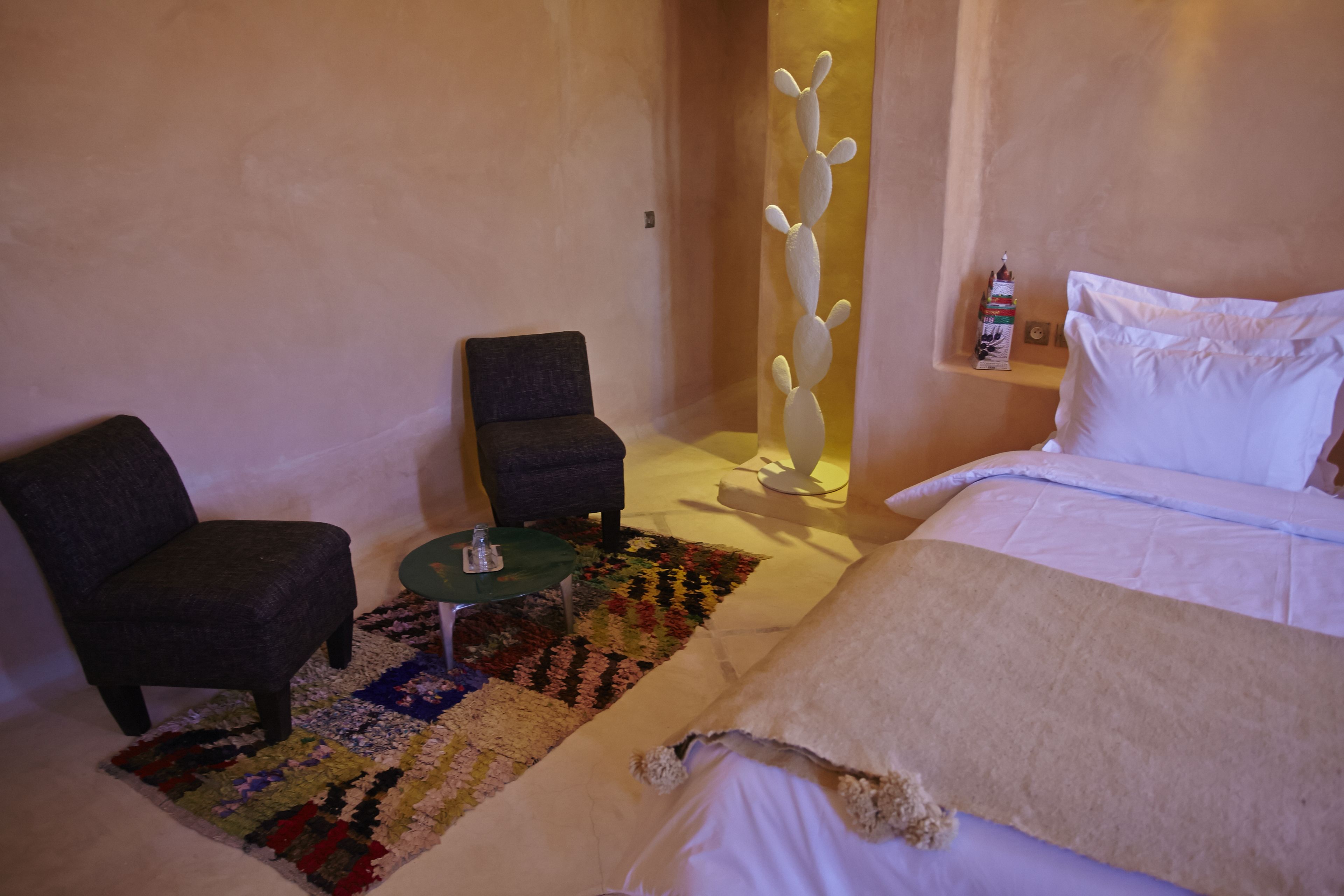 A bedroom with a bed, a chair, a small table, a colorful rug, and a cactus decoration.