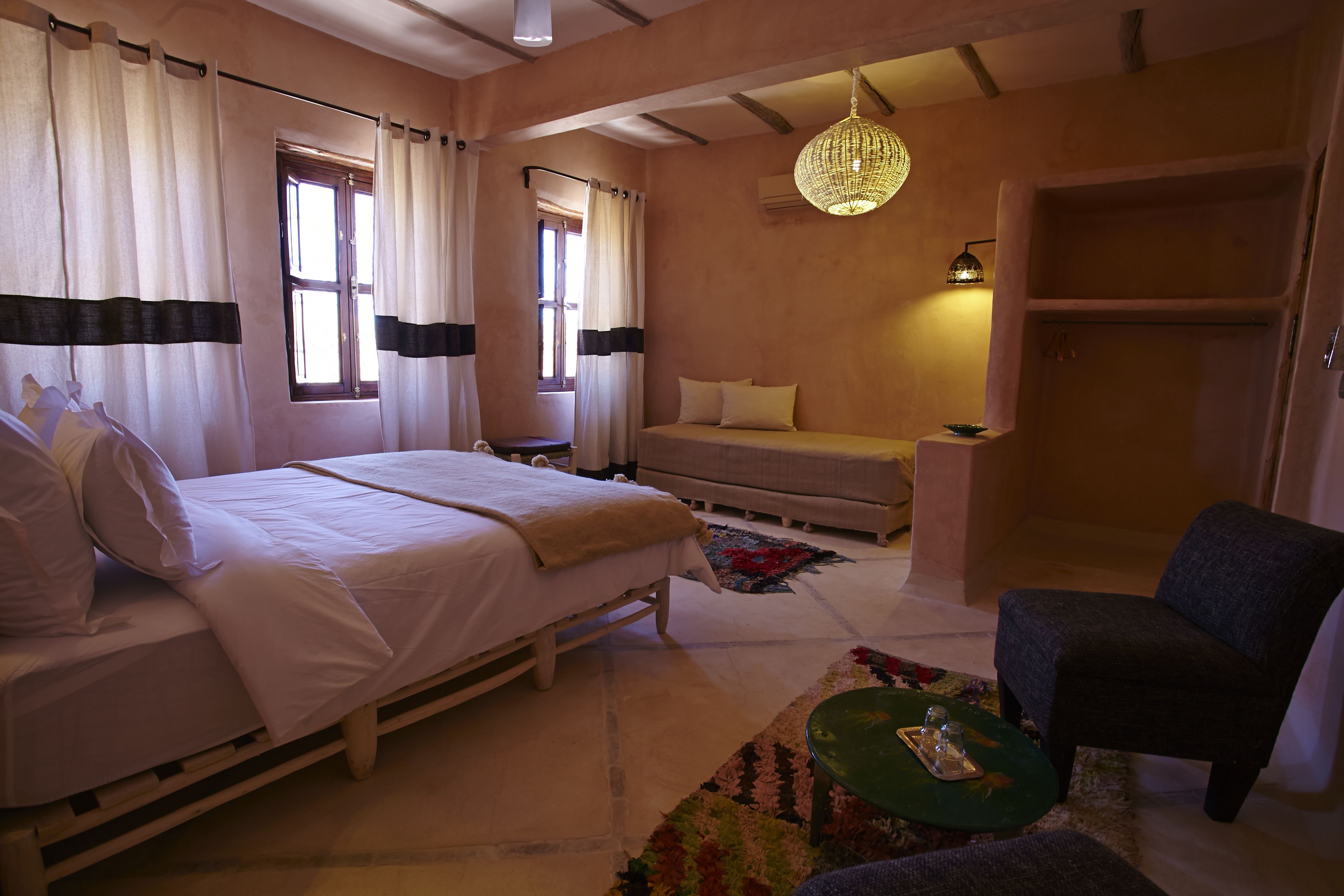 Un dormitorio con cama, sofá, sillón y una mesita. Hay cortinas en las ventanas, una lámpara colgante y un armario.