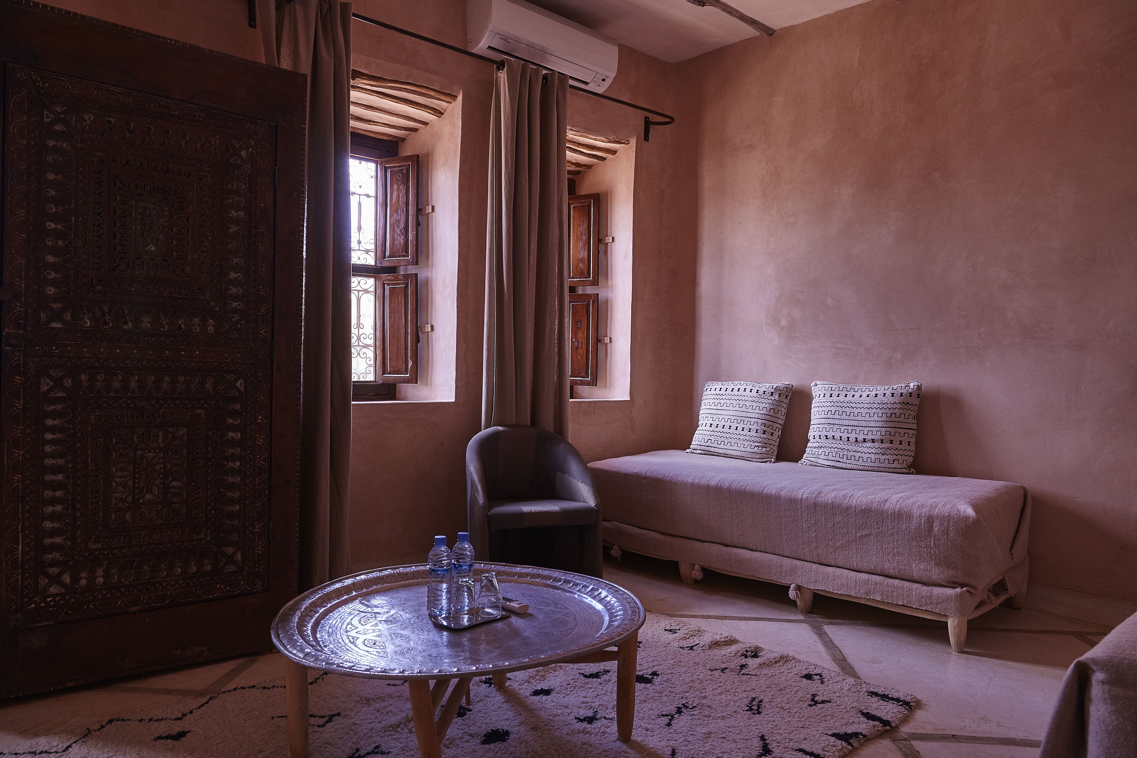 A room with a traditional wooden door, a sofa with patterned cushions, a small table with bottles, and a chair.
