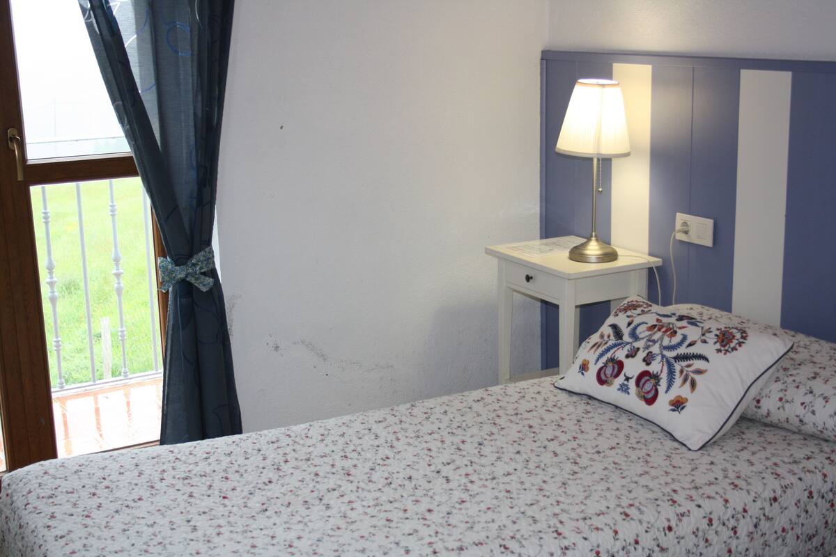 Une chambre à coucher avec un lit, une table de chevet avec une lampe, une fenêtre avec des rideaux et une vue sur l’extérieur.