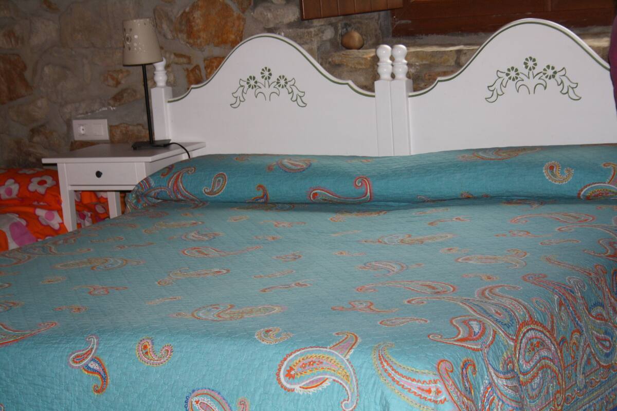 Un lit recouvert d’une couvre-lit à motifs, une table de chevet blanche avec une lampe, et un mur en pierre en arrière-plan.
