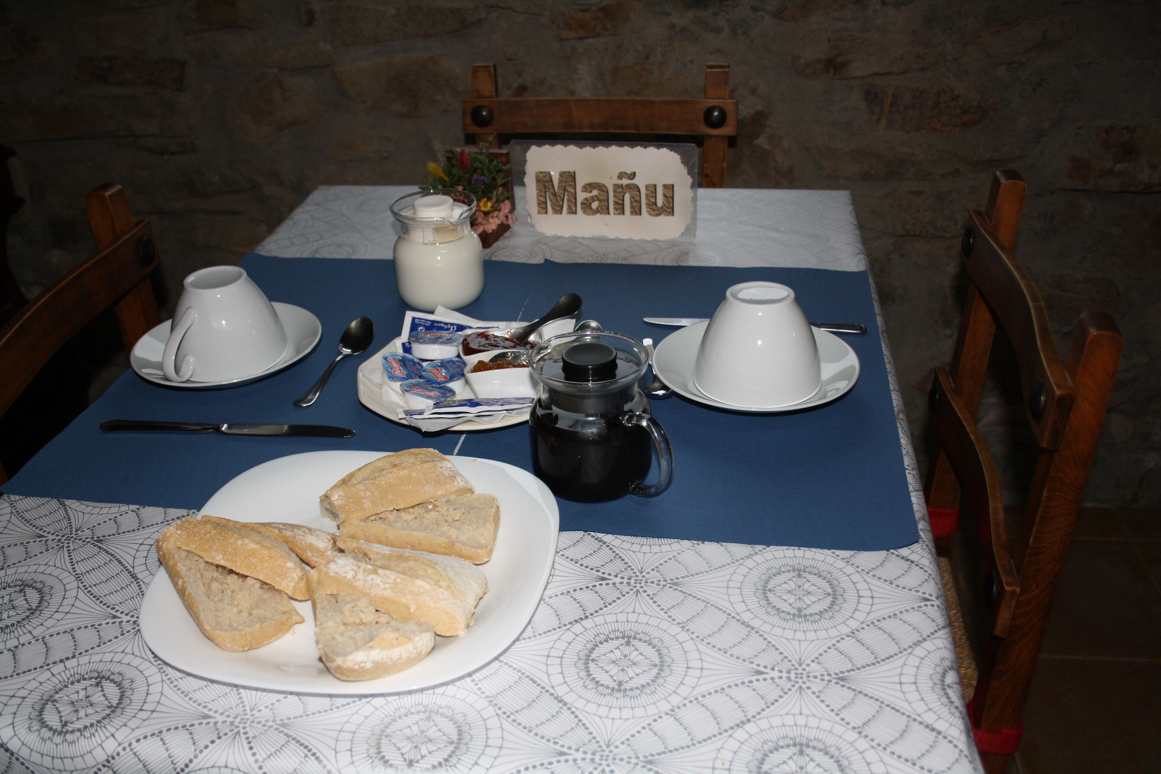 A table set with a coffee pot, cups, saucers, a plate of bread, and a sign reading "Manu".