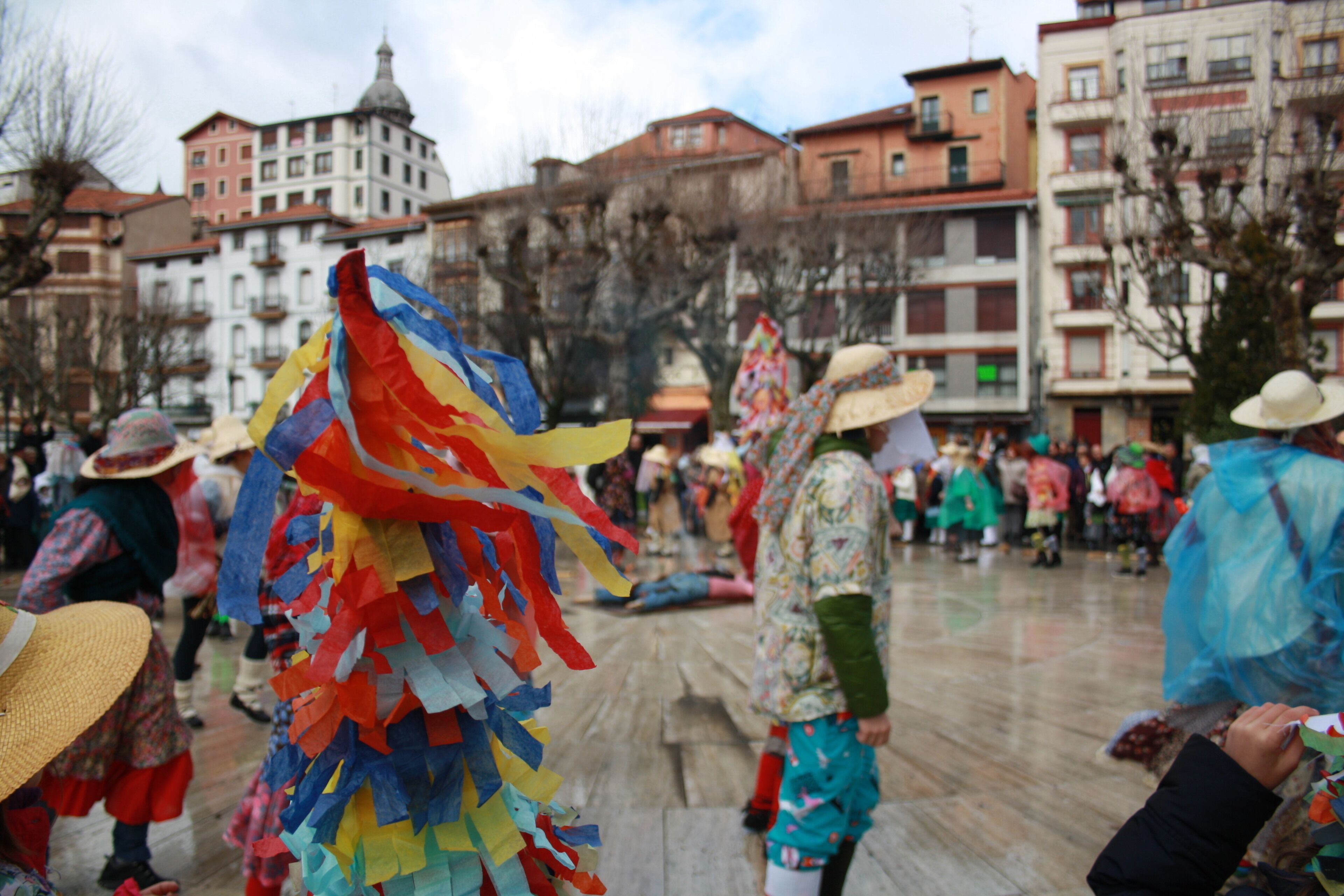 A plaza with people in colorful traditional attire, surrounded by historic buildings with balconies and a domed structure in the background.