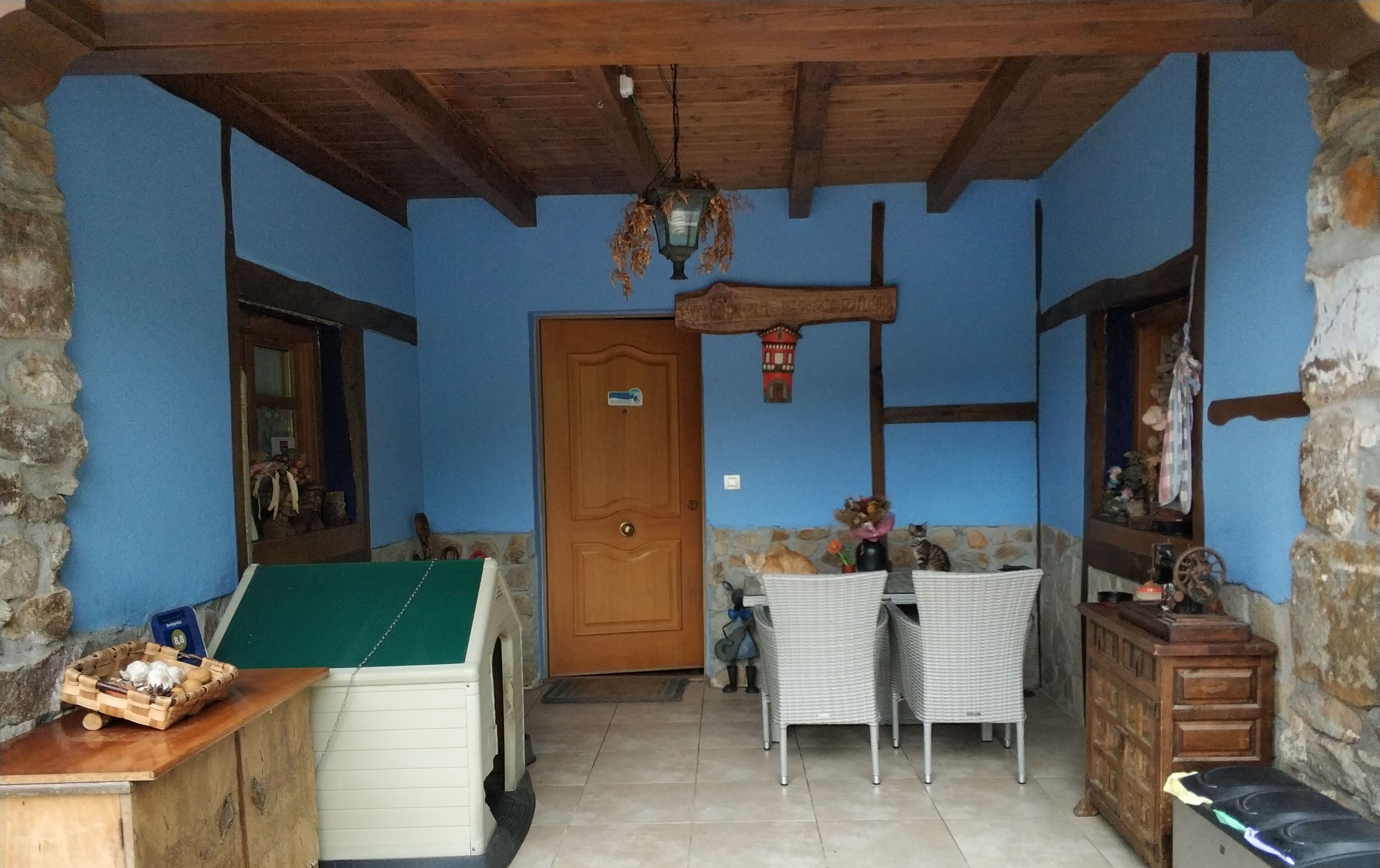 Quarto rústico com teto de madeira, parede de pedra e porta de madeira. Há uma cadeira de vime, uma mesa pequena com um cesto e um armário com espelho.