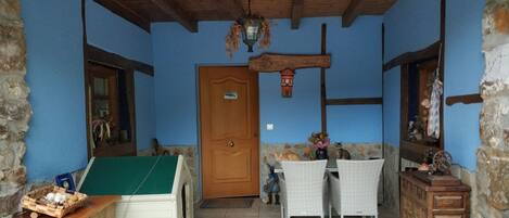 Quarto rústico com teto de madeira, parede de pedra e porta de madeira. Há uma cadeira de vime, uma mesa pequena com um cesto e um armário com espelho.