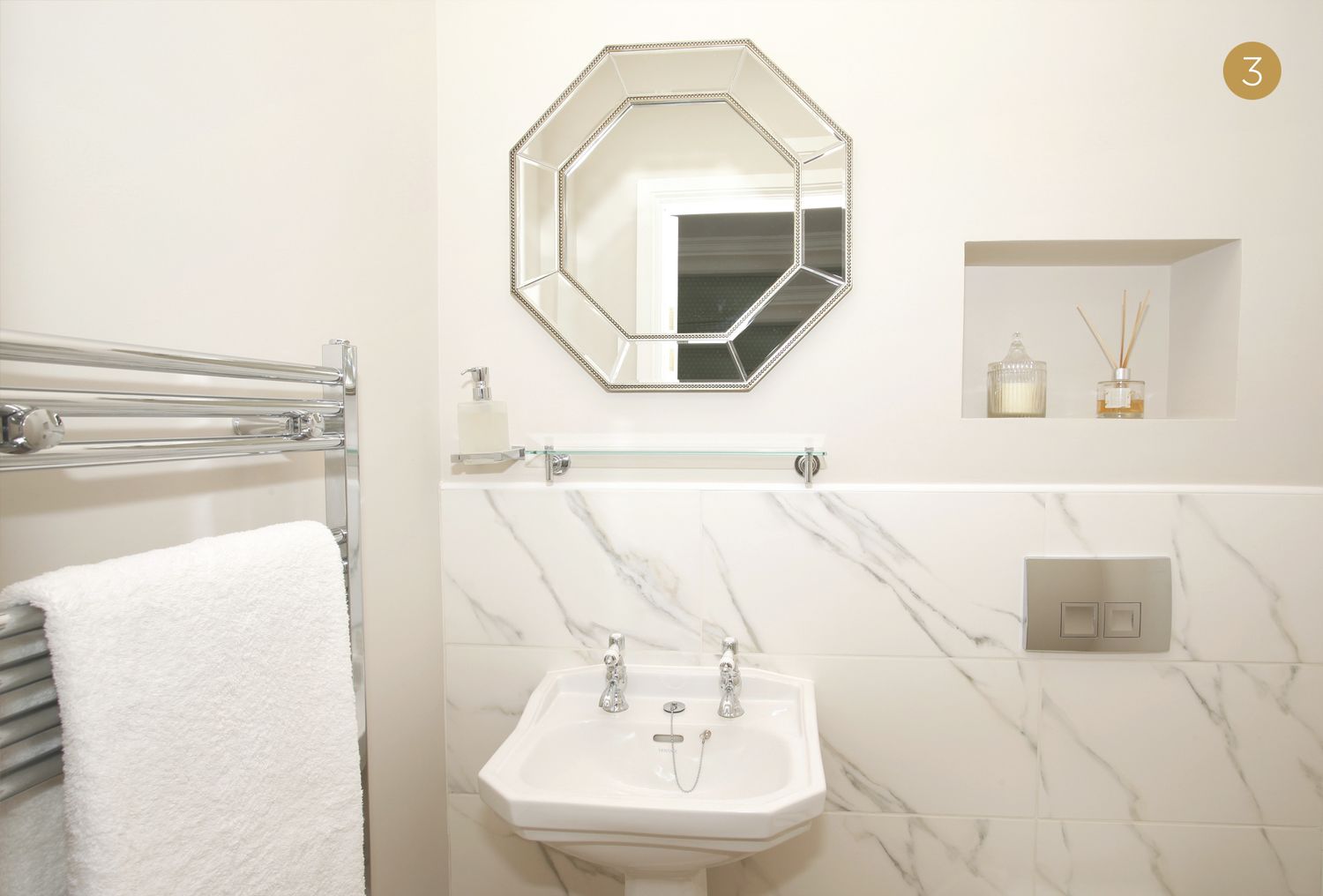 A bathroom with a white marble sink, a towel rack with a white towel, a hexagonal mirror, and a small shelf with two bottles.