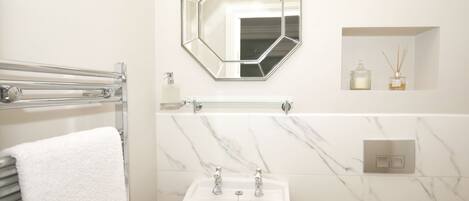 A bathroom with a white marble sink, a towel rack with a white towel, a hexagonal mirror, and a small shelf with two bottles.