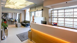 Un hall d’hôtel moderne comprenant un comptoir d’accueil, un espace salon avec un canapé, une grande fenêtre avec des stores à motifs et un luminaire au plafond.