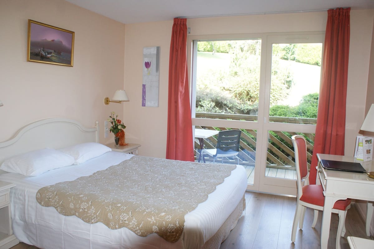 A bedroom with a bed, a desk, and a chair. There is a window with curtains and a view of greenery outside.