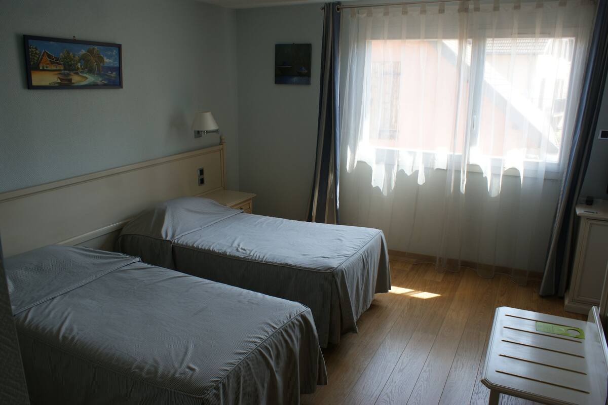 A room with two single beds, a bedside table with a lamp, a small desk, and a window with sheer curtains.
