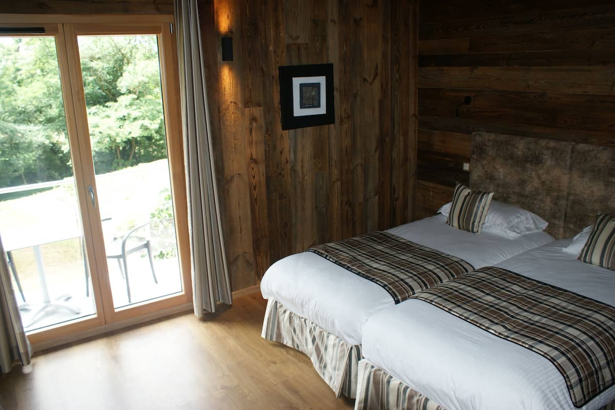 A bedroom with two beds, wooden walls, a large window, and a view of greenery outside.