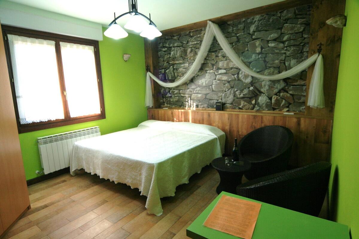 Una habitación con cama, una silla, una mesa, un muro de piedra y un ventanal con cortinas.