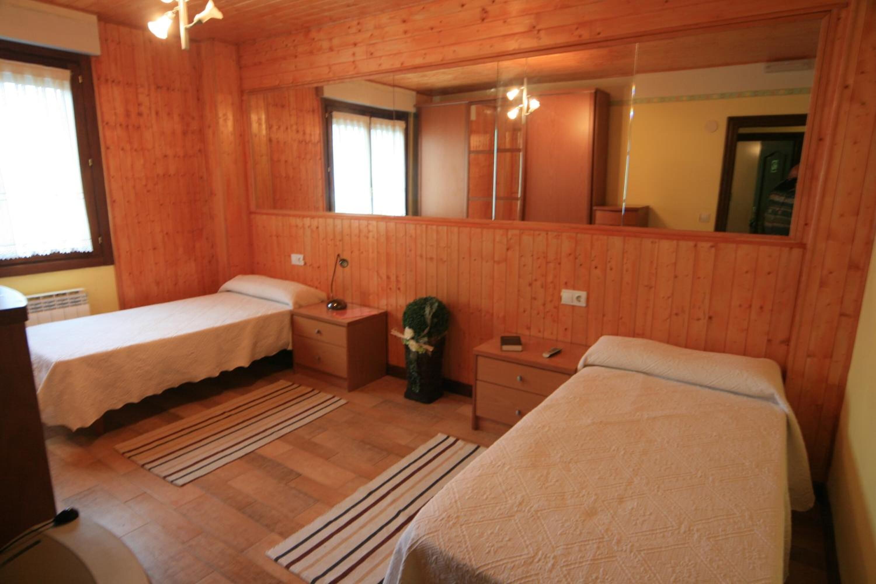Une pièce aux murs lambrissés, comprenant deux lits simples, un petit bureau, un miroir et une fenêtre avec des rideaux.