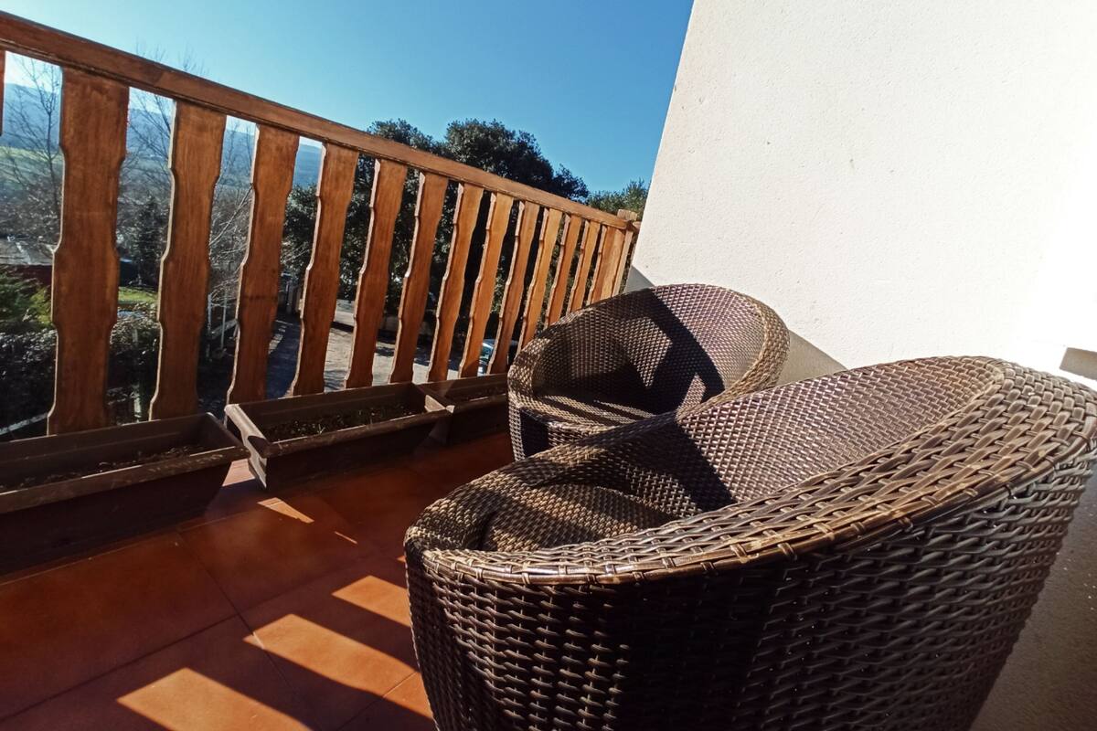 Una silla de mimbre en un balcón con baranda de madera, con vistas a un paisaje de árboles y cielo despejado.