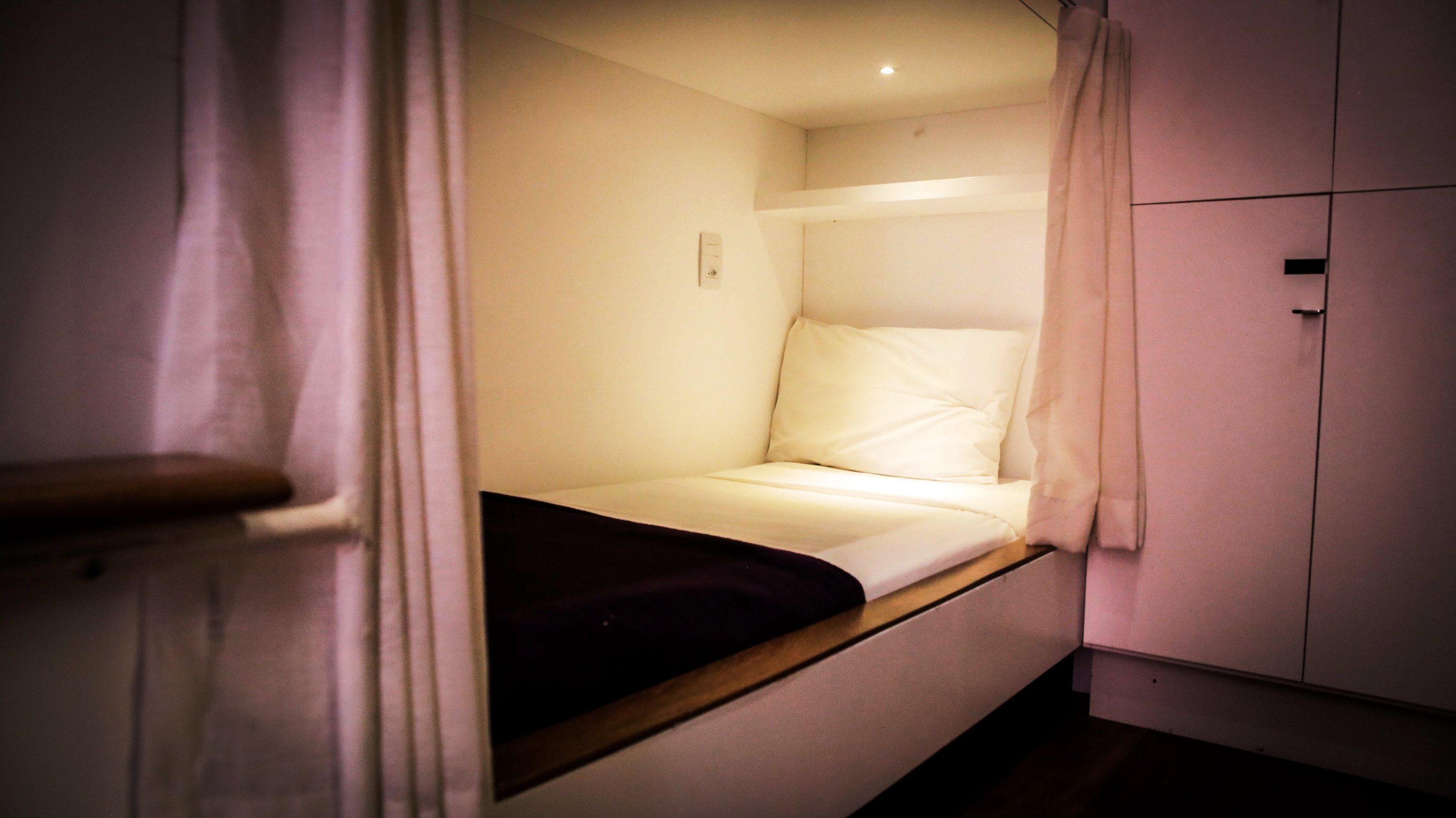 Una habitación pequeña con cama individual, cabecera de madera, colchón blanco y manta oscura.