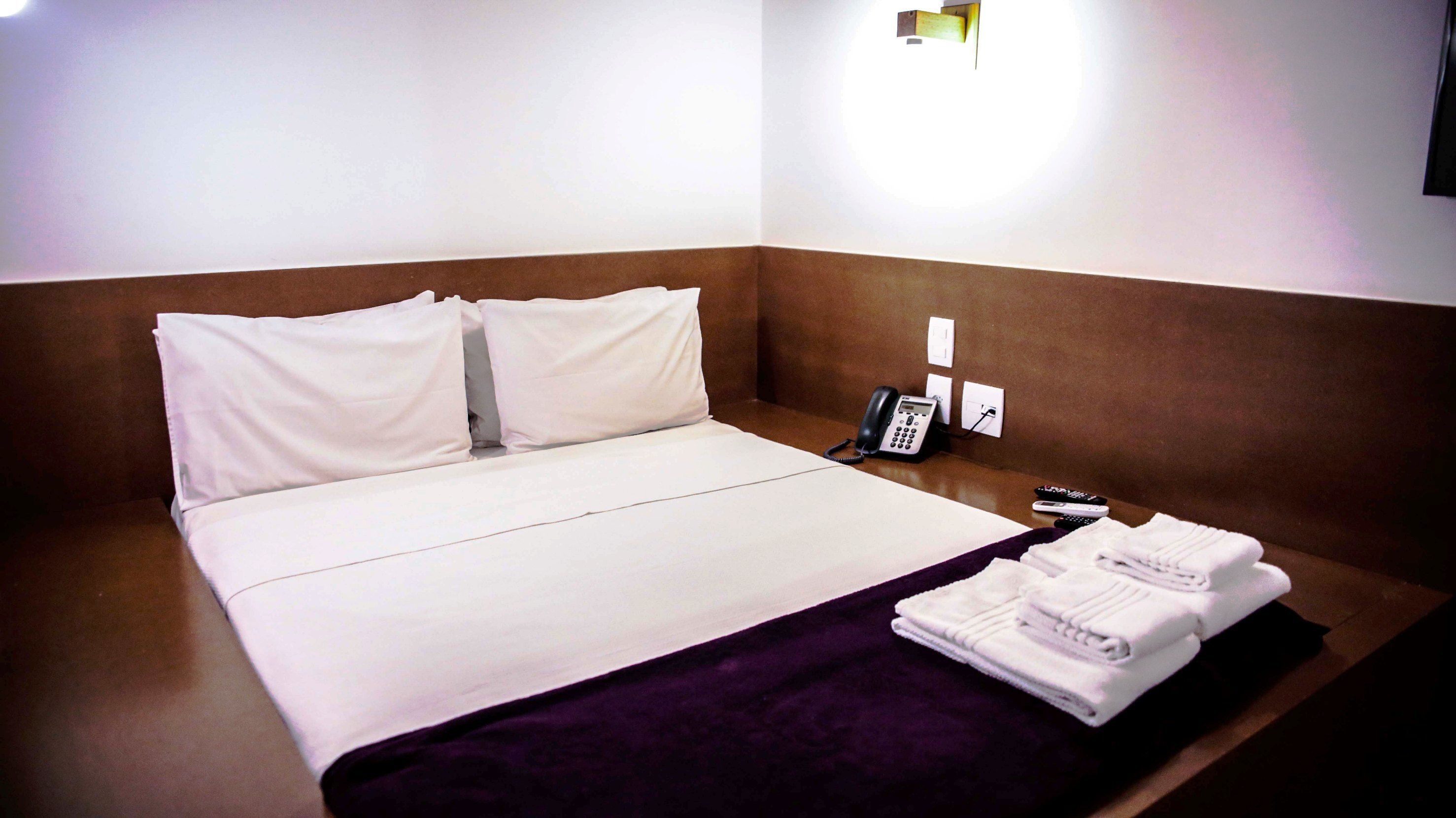 A hotel room with a neatly made bed, a bedside phone, and a set of towels on a tray.