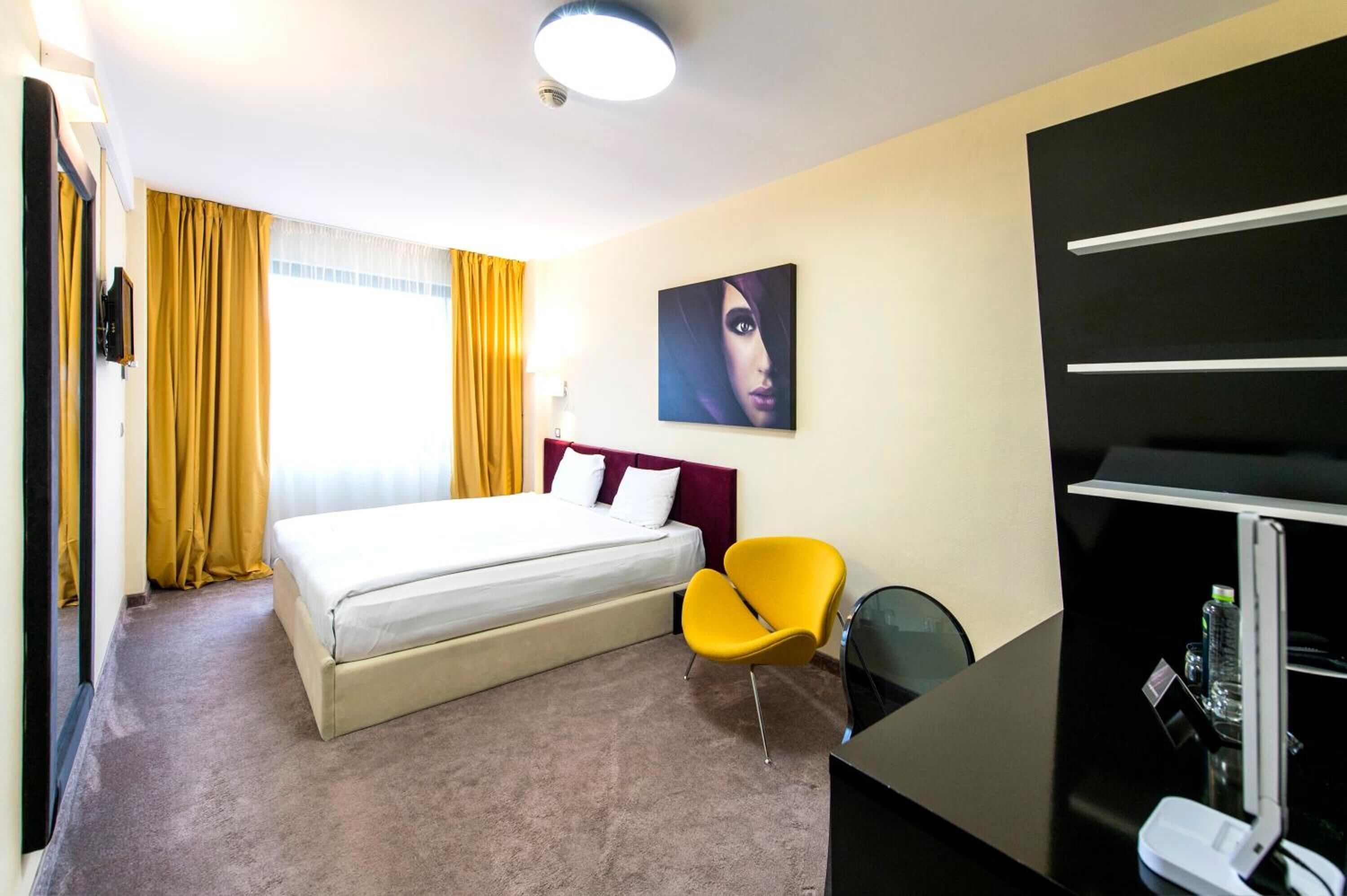 A hotel room with a bed, a yellow chair, a desk with a lamp, and a painting on the wall.