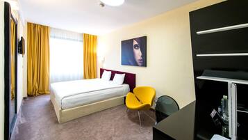 A hotel room with a bed, a yellow chair, a desk with a lamp, and a painting on the wall.