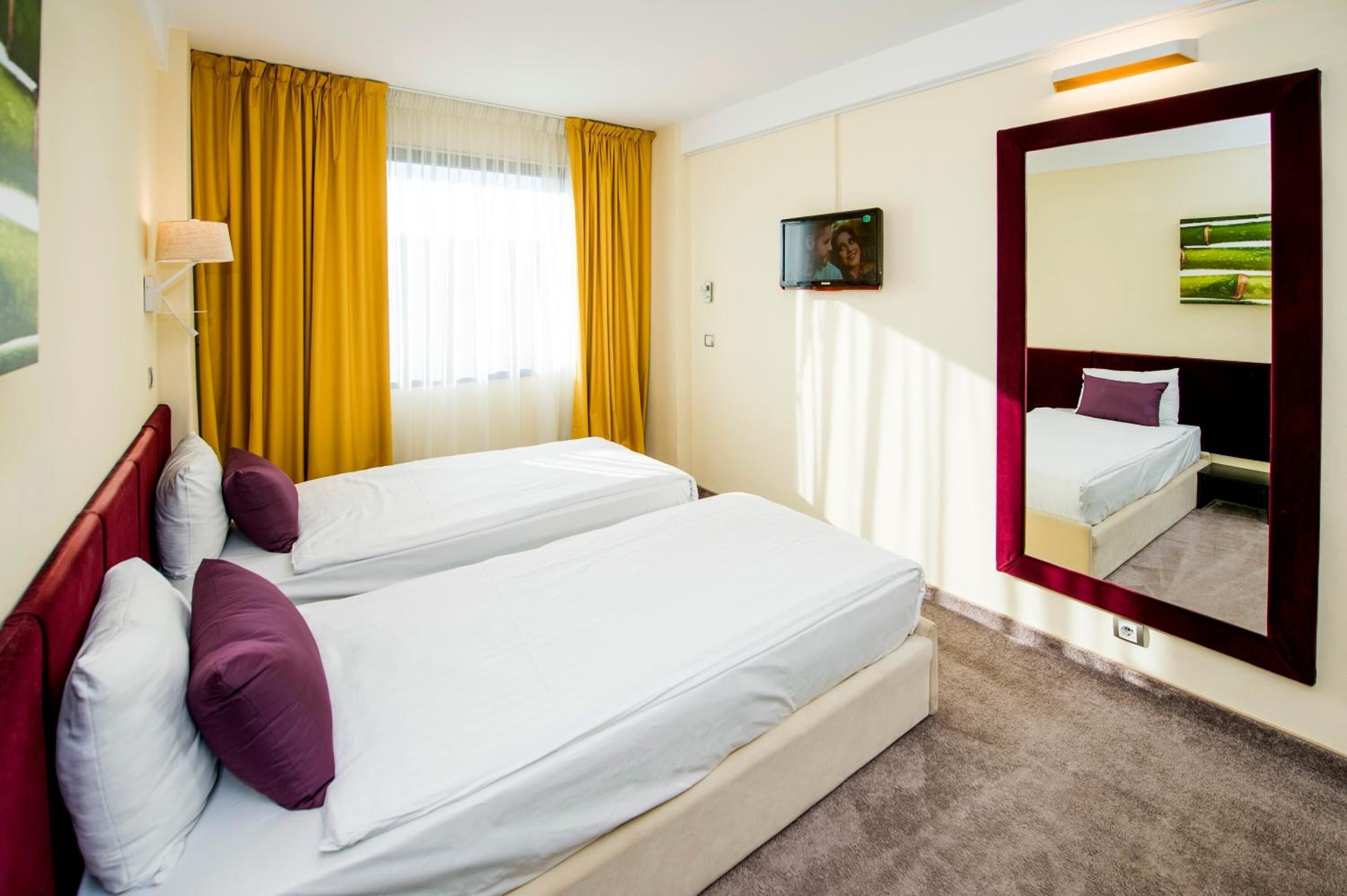 A hotel room with two beds, a large mirror, a TV, and a window with yellow curtains.