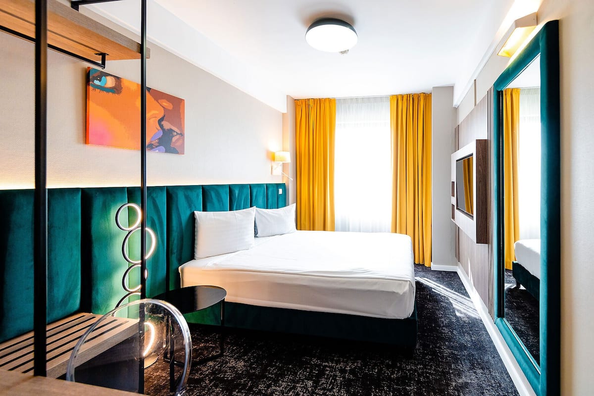 A hotel room with a bed, a desk, a chair, a mirror, and a painting on the wall.