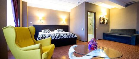Una habitación de hotel con una cama, un sillón amarillo, un sofá azul y una mesa de centro de vidrio.