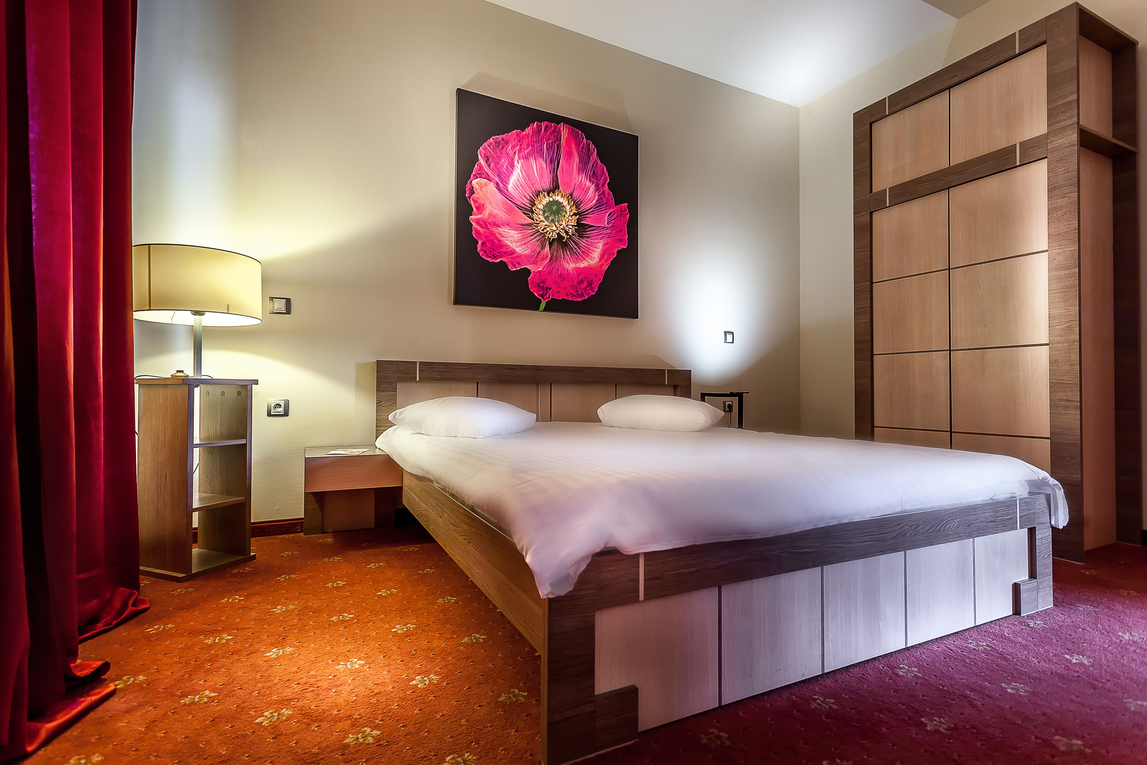 A hotel room with a wooden bed, a large floral artwork on the wall, a red carpet, and a wooden wardrobe.