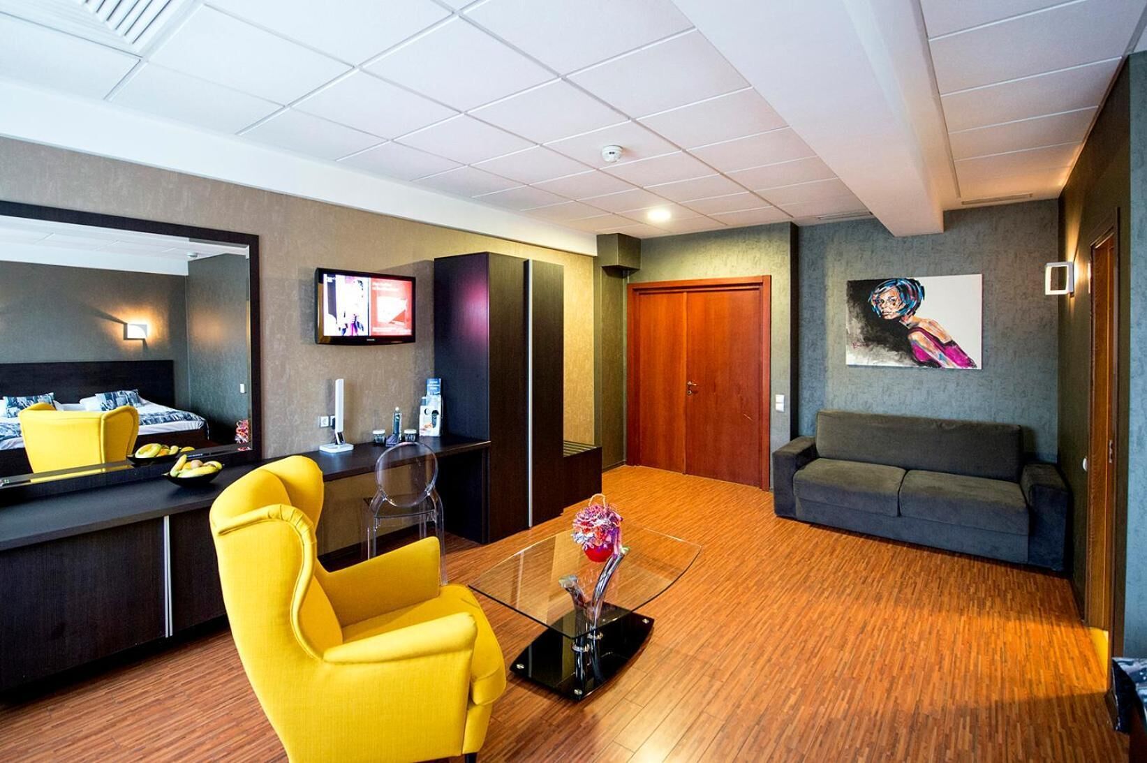 A modern hotel lobby with a reception desk, a yellow armchair, a sofa, a wooden floor, and a painting on the wall.