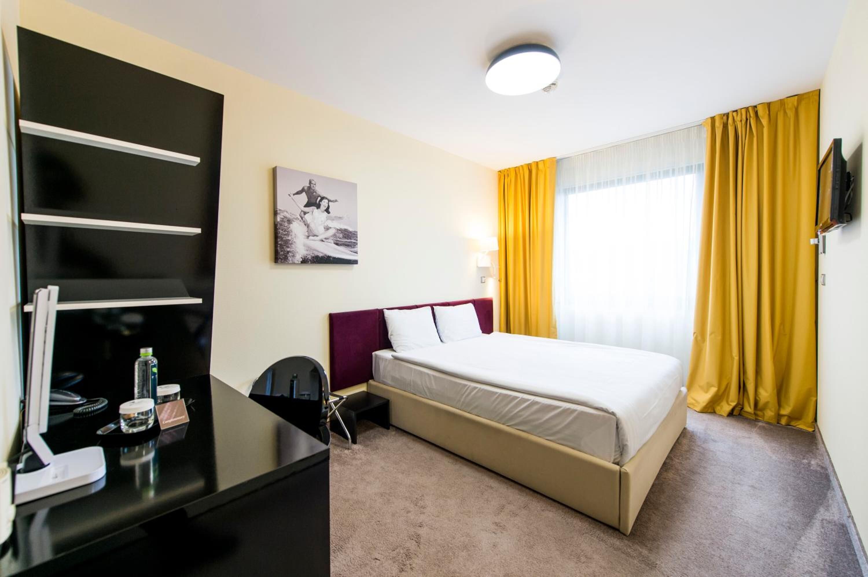 A hotel room with a bed, a desk with a computer, a chair, a desk lamp, and a window with yellow curtains.